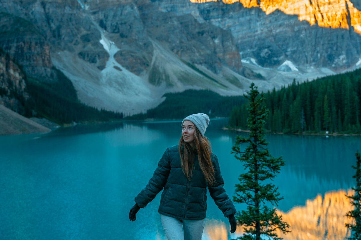 girl looking to the side in a green jacket and grey toque in front of lake with mountains in the background