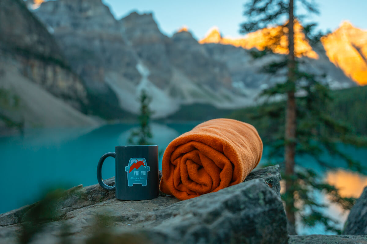 Via Via Moraine Lake mug and orange blanket sitting on a rock with the lake and mountains in the. background